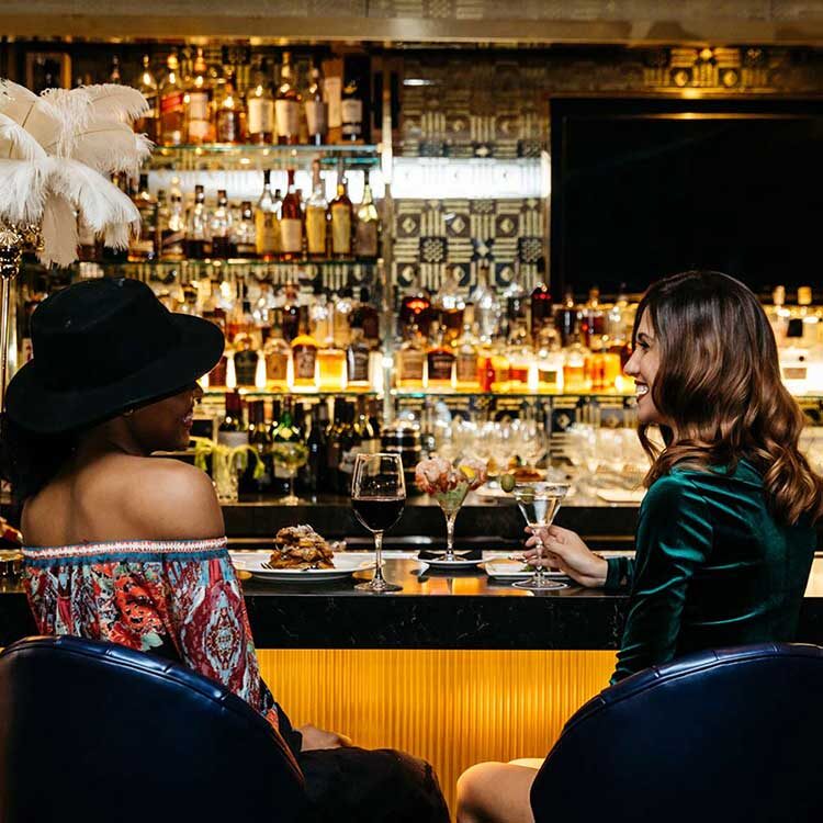 two women sitting at a bar smiling at each other with cocktails in hand